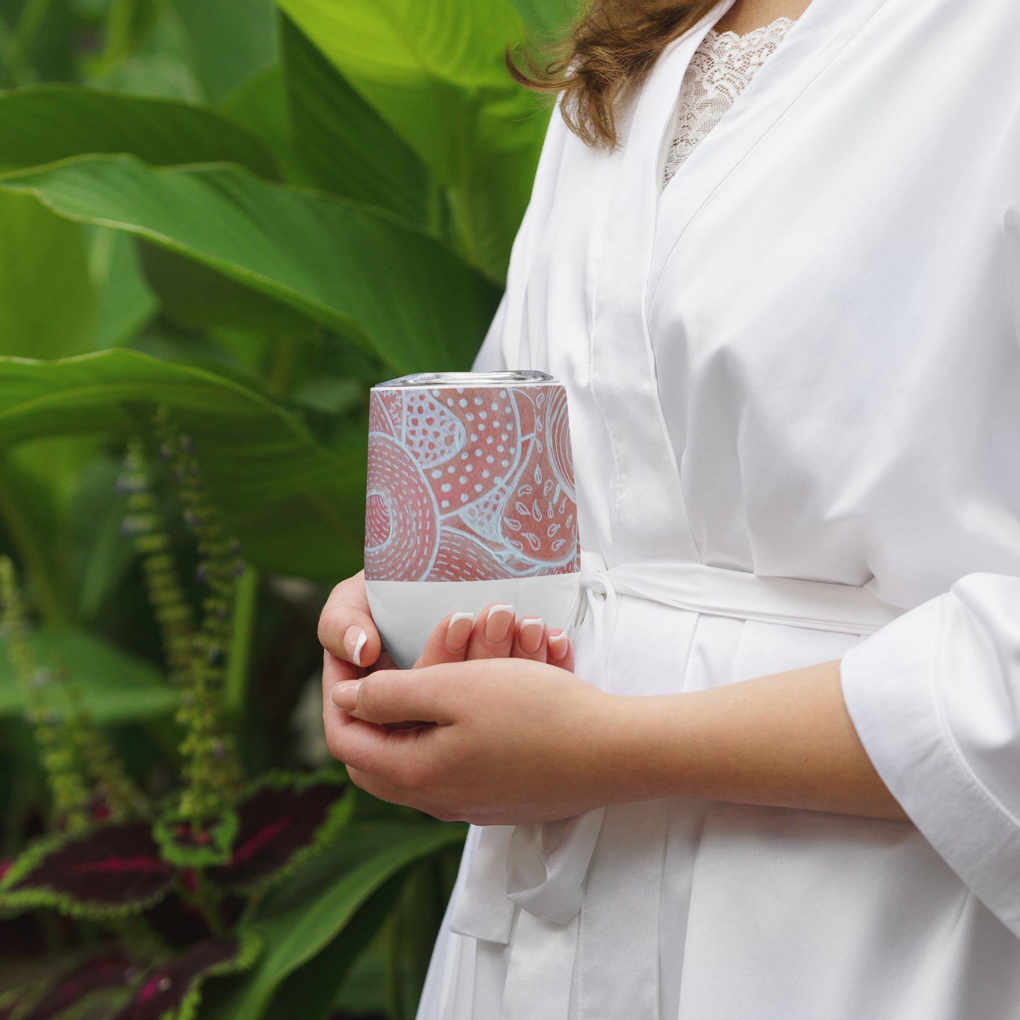 Stainless Steel; wine tumbler in white. Featuring beautifully designed Aboriginal artwork from the Dreaming Series in Pink and White. Front View. being cupped in two hands by woman in a white shirt.