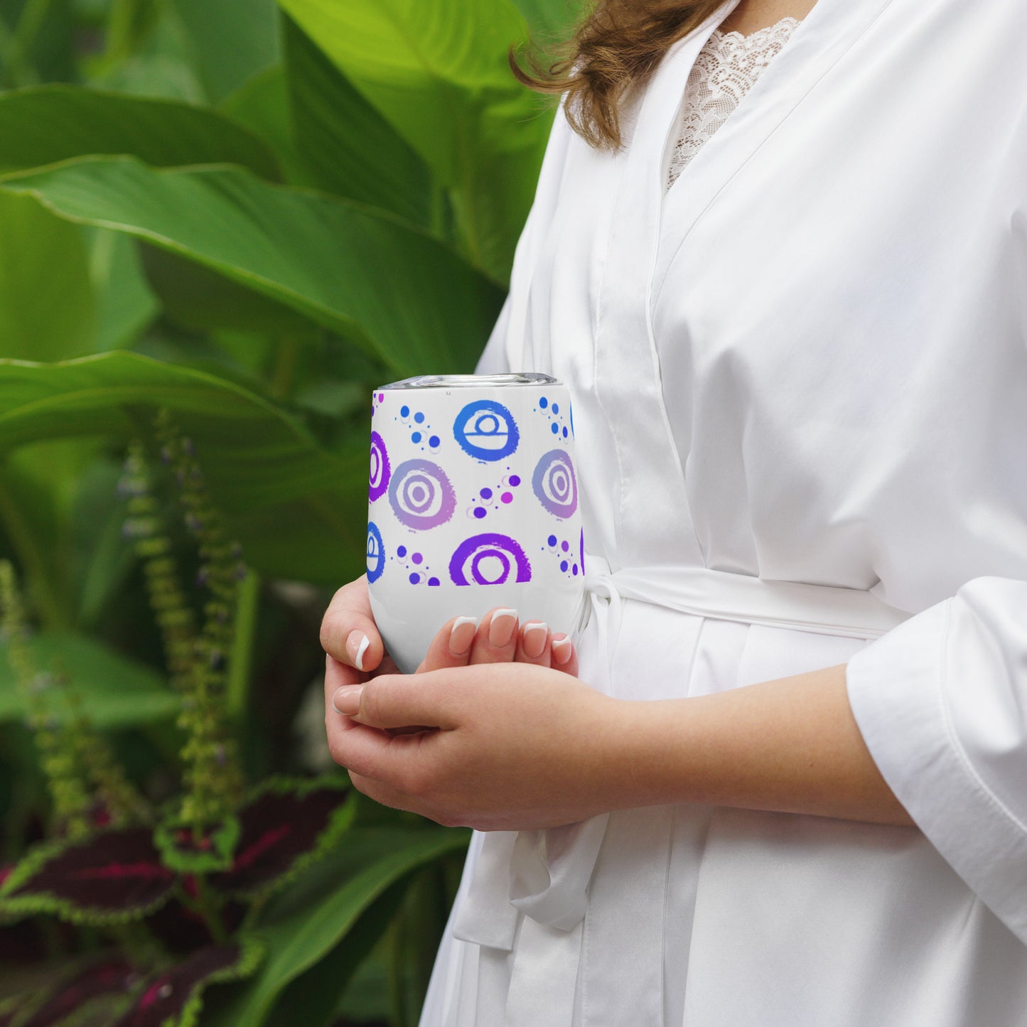 Wine tumbler in white with lid, featuring Blue, pink and Purple petroglyph designs. Front View. Being cupped in two hands by woman.
