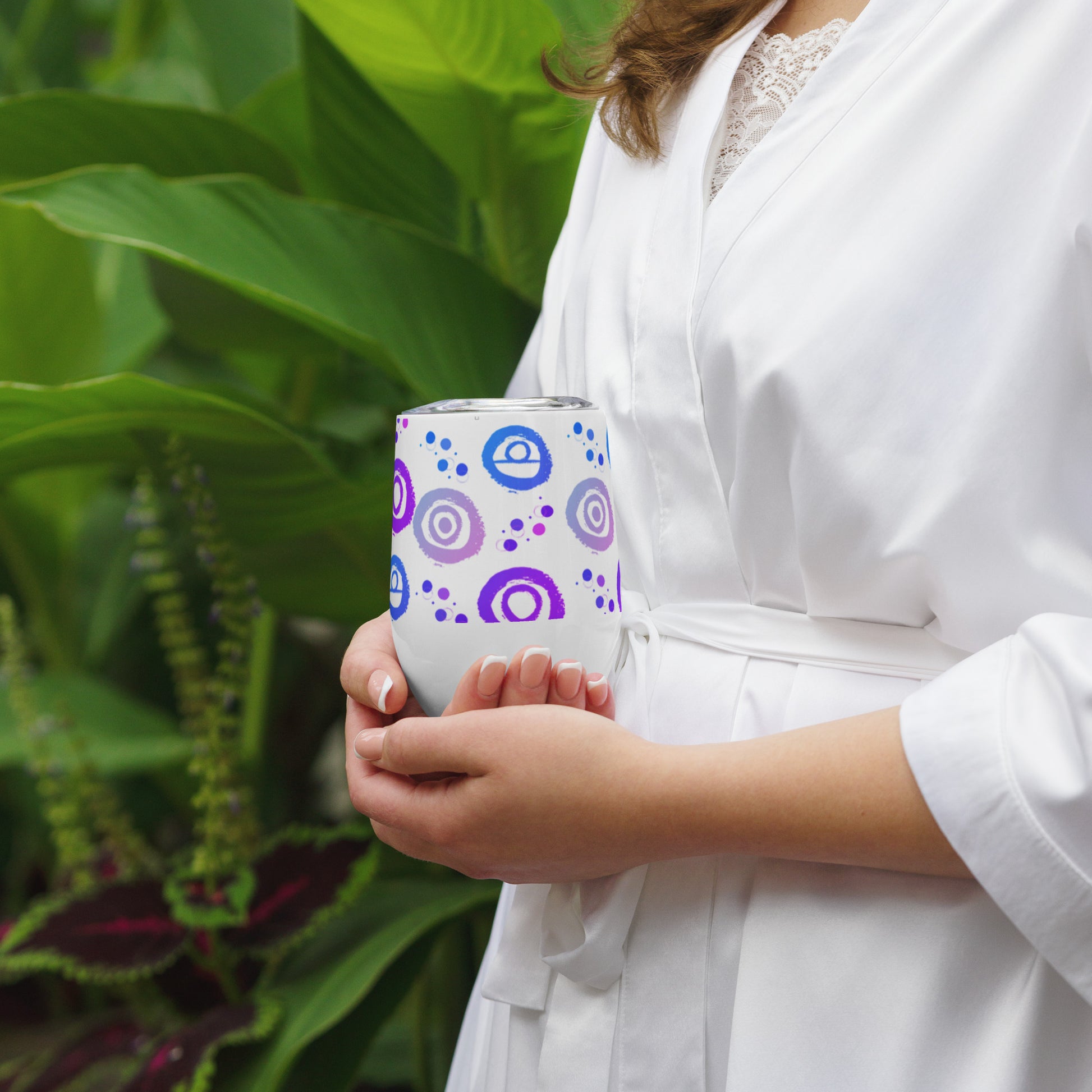 Wine tumbler in white with lid, featuring Blue, pink and Purple petroglyph designs. Front View. Being cupped in two hands by woman.