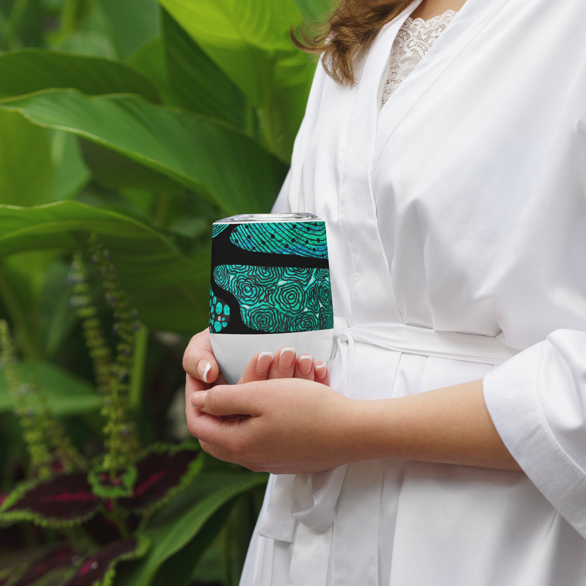 Wine tumbler in White with lid featuring beautiful black, green and white Aboriginal Artwork. Front View. Woman holding cupped in two hands.